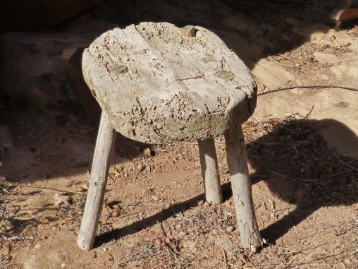 Tamboret de fusta antiga amb tres potes, desgastat pel temps i l'ús, fotografiat a l'exterior sobre terra seca. Símbol dels tres pilars del Pla de Lectura de Centre: Saber llegir, Llegir per aprendre i El gust per la lectura.