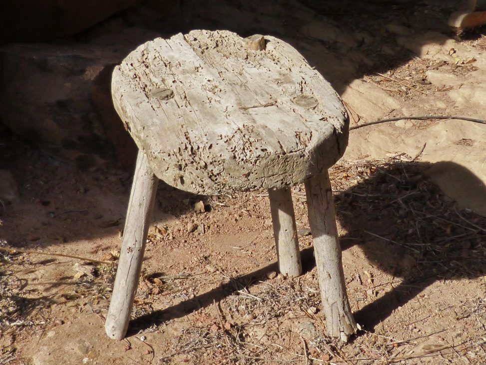 Tamboret de fusta antiga amb tres potes, desgastat pel temps i l'ús, fotografiat a l'exterior sobre terra seca. Símbol dels tres pilars del Pla de Lectura de Centre: Saber llegir, Llegir per aprendre i El gust per la lectura.